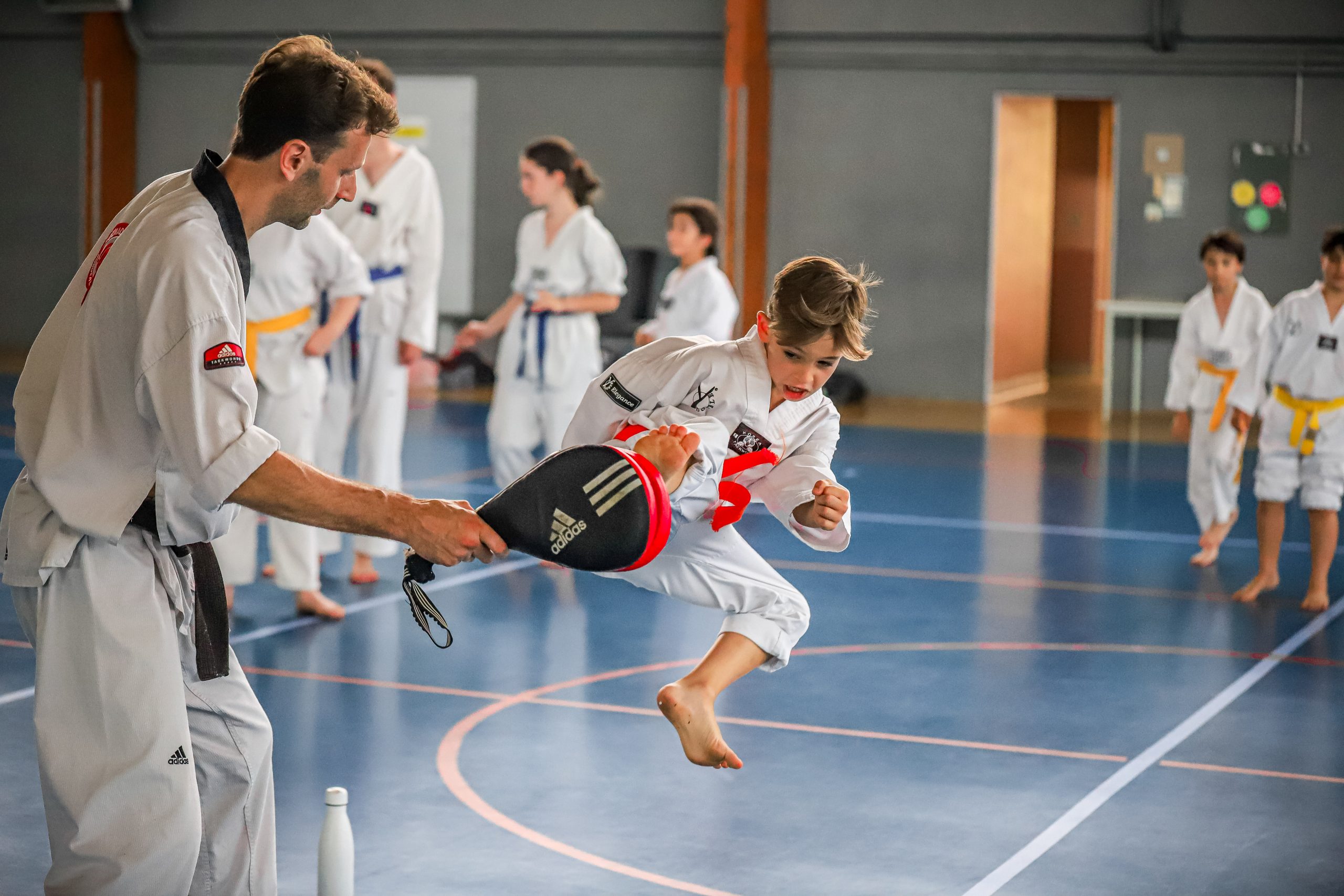Un enfant donne un coup de pied sur un raquette tenue par un enseignant dans un gymnase.